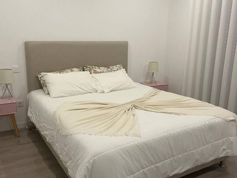 A neatly made bed with a beige headboard, white bedding, and a decorative throw. There are two bedside tables with lamps and a nightstand with a lamp.