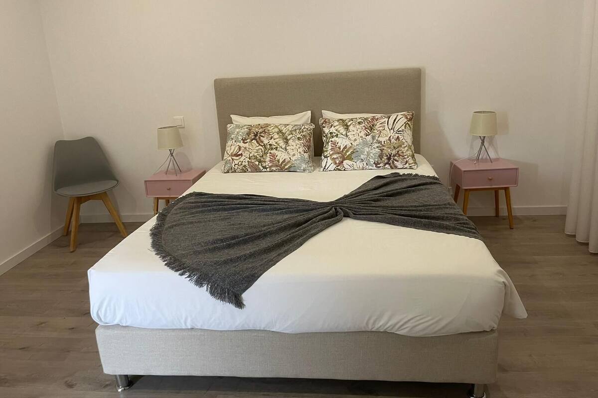 A neatly made bed with a grey throw, two floral pillows, and a headboard. There are two bedside tables with lamps and a chair.