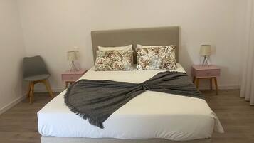 Una cama bien hecha con un plaid gris, dos almohadas florales y un cabecero. Hay dos mesitas de noche con lámparas y una silla.