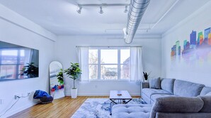 A modern living room with a sofa, a coffee table, a potted plant, a cityscape mural, and a large window. - Modern Spacious Loft in Downtown w/ free parking (Memphis)