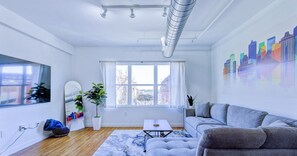 A modern living room with a sofa, a coffee table, a potted plant, a cityscape mural, and a large window.