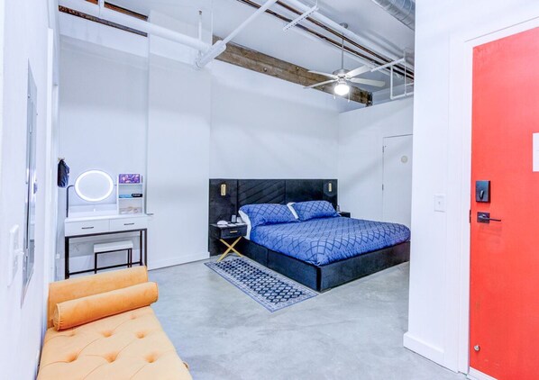 A bedroom with a bed, a bedside table, a chair, a desk with a chair, a red door, and exposed ductwork on the ceiling.