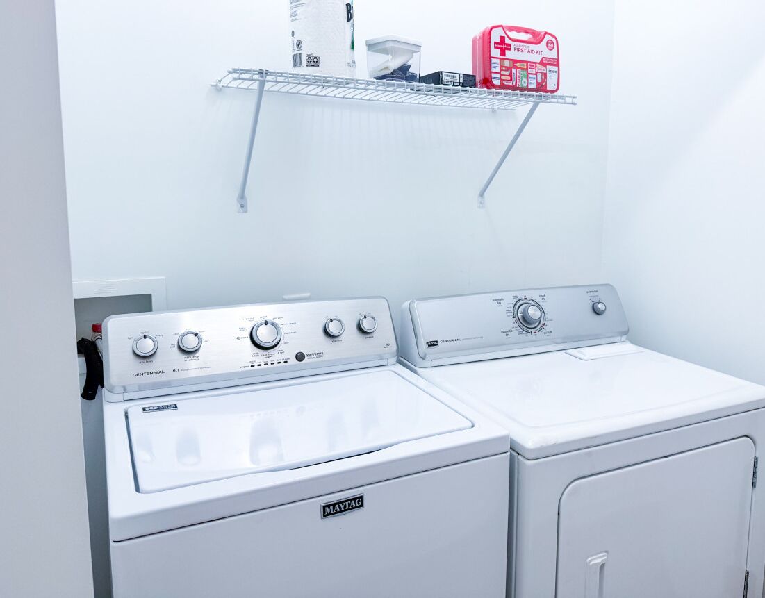 A laundry area with two Maytag washers and dryers, a metal shelf with a red first aid kit, and a white wall.