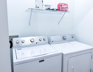 A laundry area with two Maytag washers and dryers, a metal shelf with a red first aid kit, and a white wall.