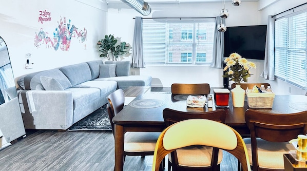 Ein modernes Wohnzimmer mit einem Ecksofa, einem Esstisch, Stühlen, einem Flachbildfernseher und einem großen Fenster mit Jalousien.