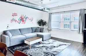 Ein modernes Wohnzimmer mit einem grauen Ecksofa, einem Holztisch, einem gemusterten Teppich, einem großen Fenster mit Jalousien und einer Stadtbild-Wandkunst.