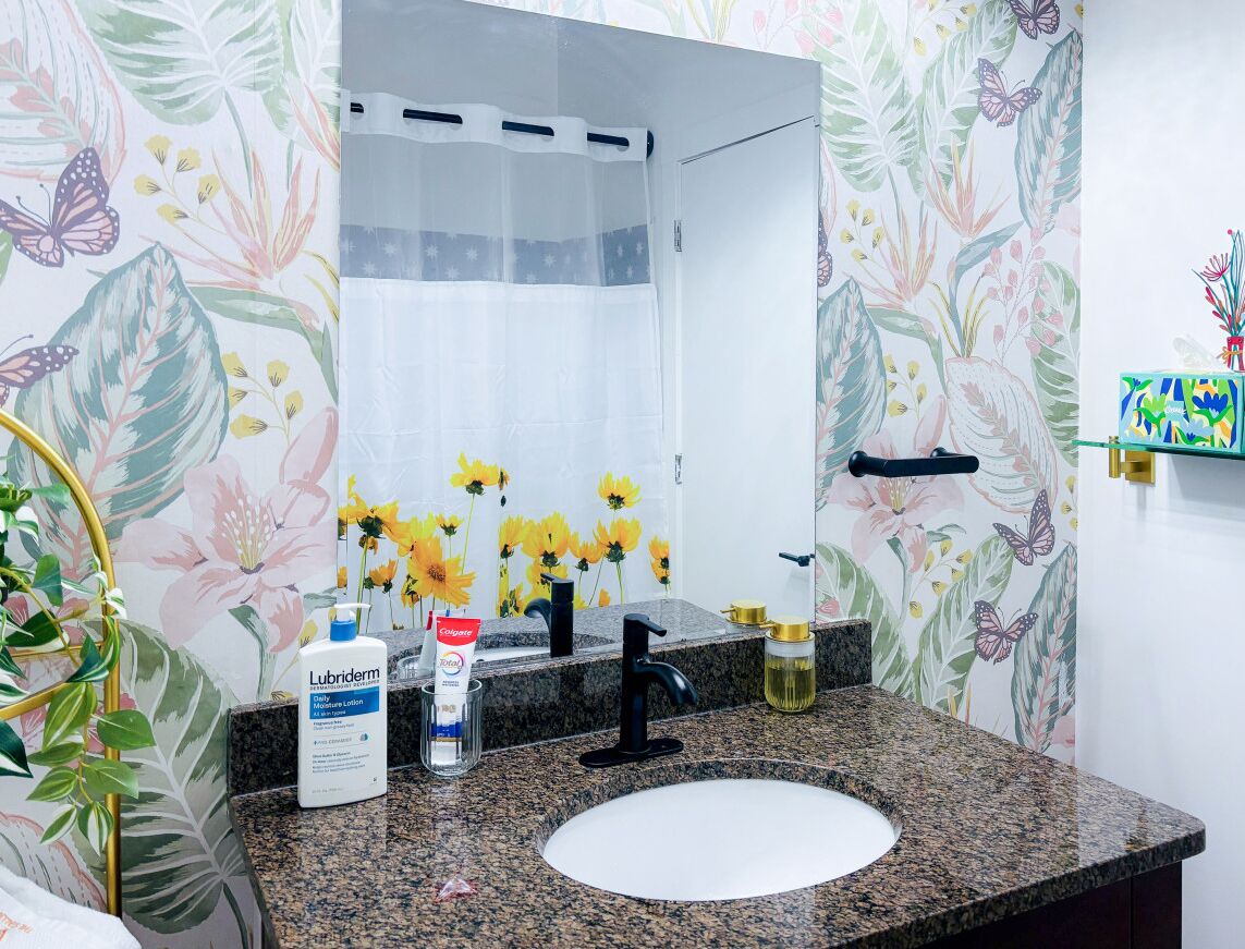 A bathroom with a granite countertop, a white sink, a black faucet, a mirror, and floral wallpaper.