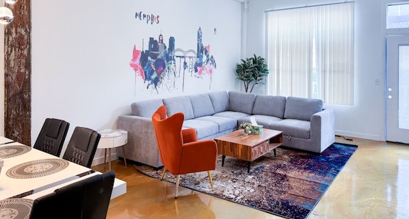 Un salon moderne avec un canapé d’angle gris, une table basse en bois et un tapis aux couleurs vives.