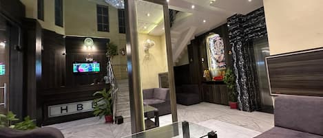 A modern lobby with a glass coffee table, dark sofas, a reception desk, and a large mirror reflecting the space.