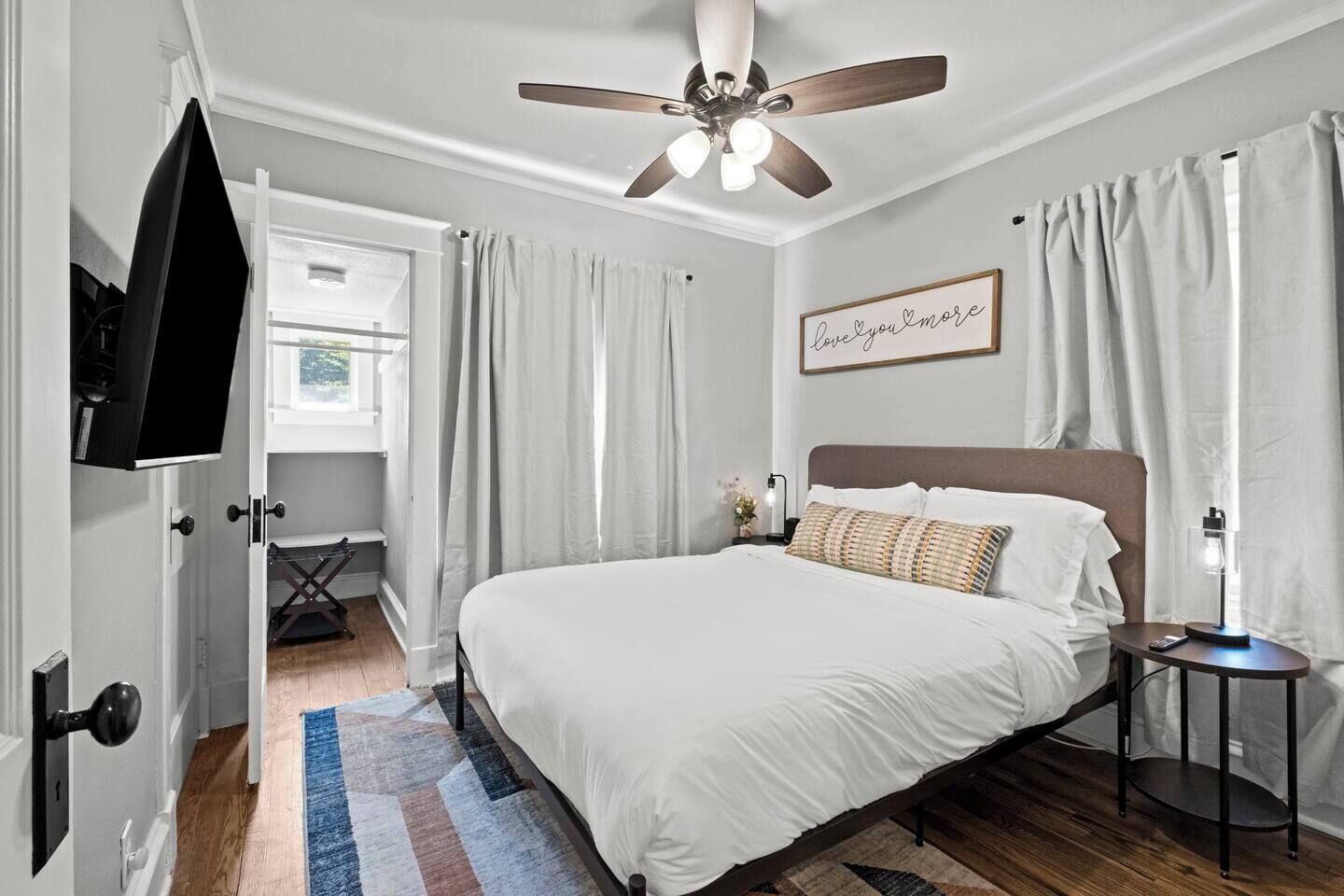Een slaapkamer met een bed, een plafondventilator, een nachtkastje, een aan de muur bevestigde televisie en een raam met gordijnen.