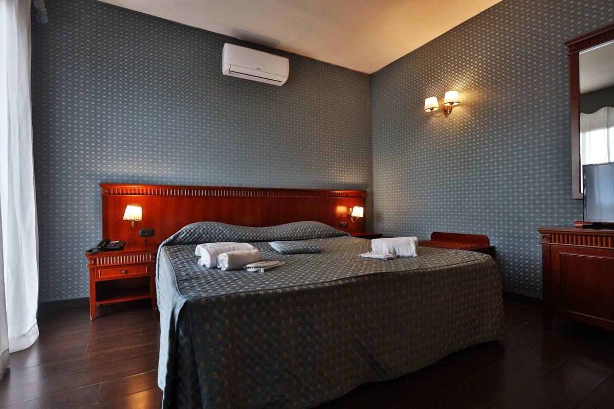 A hotel room with a large bed, two bedside tables with lamps, a wooden headboard, an air conditioning unit, a television, and a mirror.
