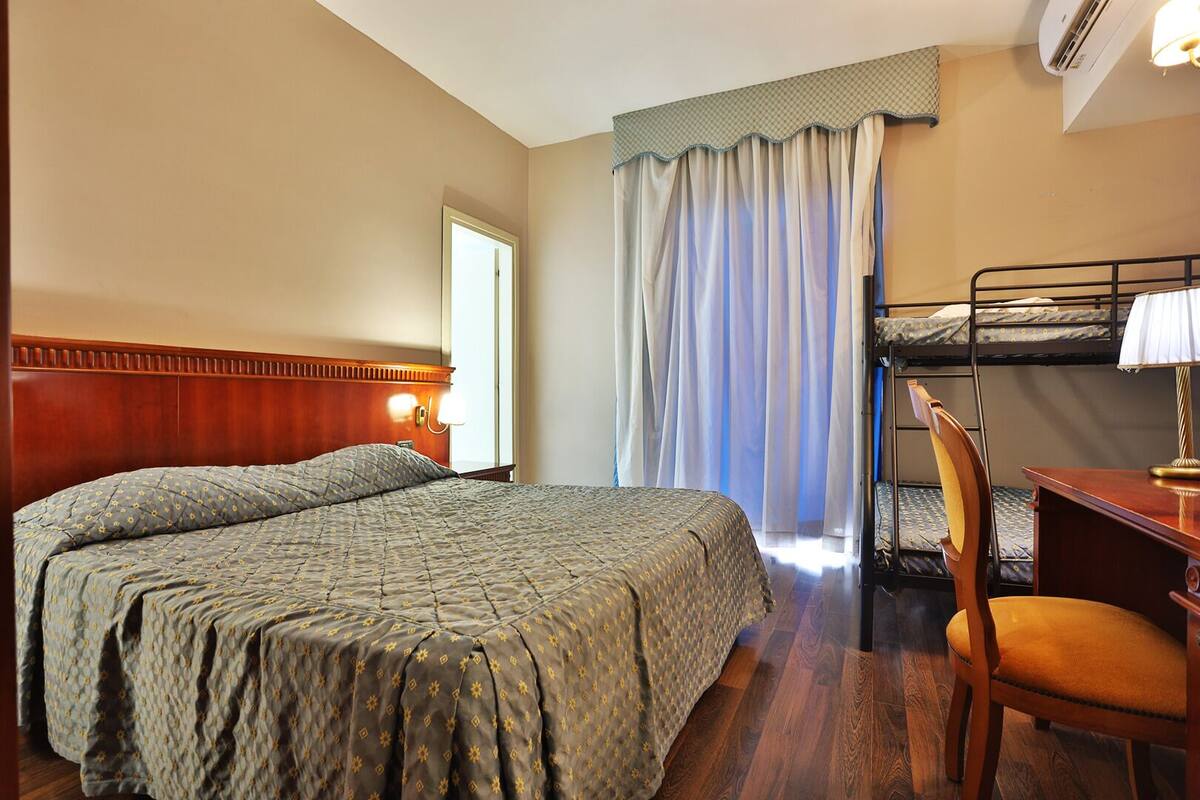 A hotel room with a double bed, a desk with a chair, a bunk bed, and a window with curtains.