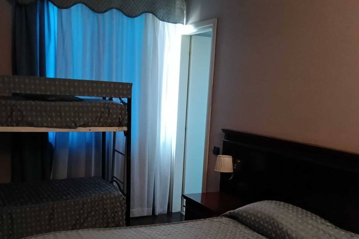 A hotel room with a bunk bed, a single bed, a nightstand, a lamp, and a view of the outside through a window with curtains.