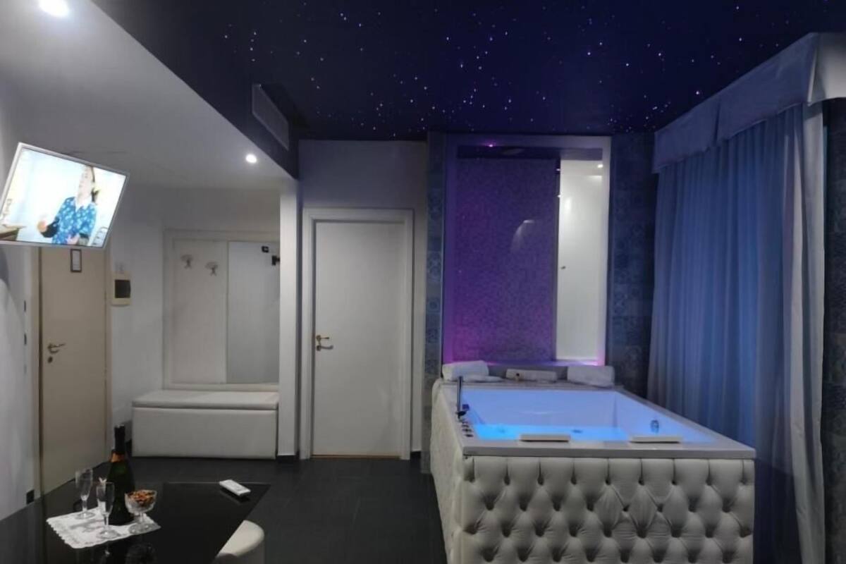 A bedroom with a starry ceiling, a bed with a tufted headboard, a small table with a bottle and glasses, and a TV on the wall.