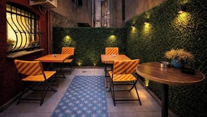 Uma área externa com mesas e cadeiras, um tapete azul estampado e uma parede verde com plantas.