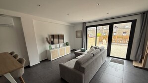 A modern living room with a grey sectional sofa, a dining table with chairs, a white console table with a TV, and a large sliding glass door leading to an outdoor area. - Gîte TI BOEM, near Cancale, Saint-Malo and Mont-Saint-Michel (La Fresnais)