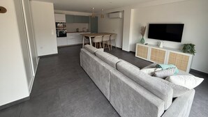 A modern living room with a grey sectional sofa, a dining area with a wooden table and chairs, a kitchen in the background, and a TV mounted on the wall. - Gîte TI BOEM, near Cancale, Saint-Malo and Mont-Saint-Michel (La Fresnais)