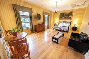 Habitación de hotel con piso de madera, cama con estructura metálica, mesa de comedor con sillas, un pequeño banco, un sofá, un televisor, un cuadro y una ventana con cortinas.