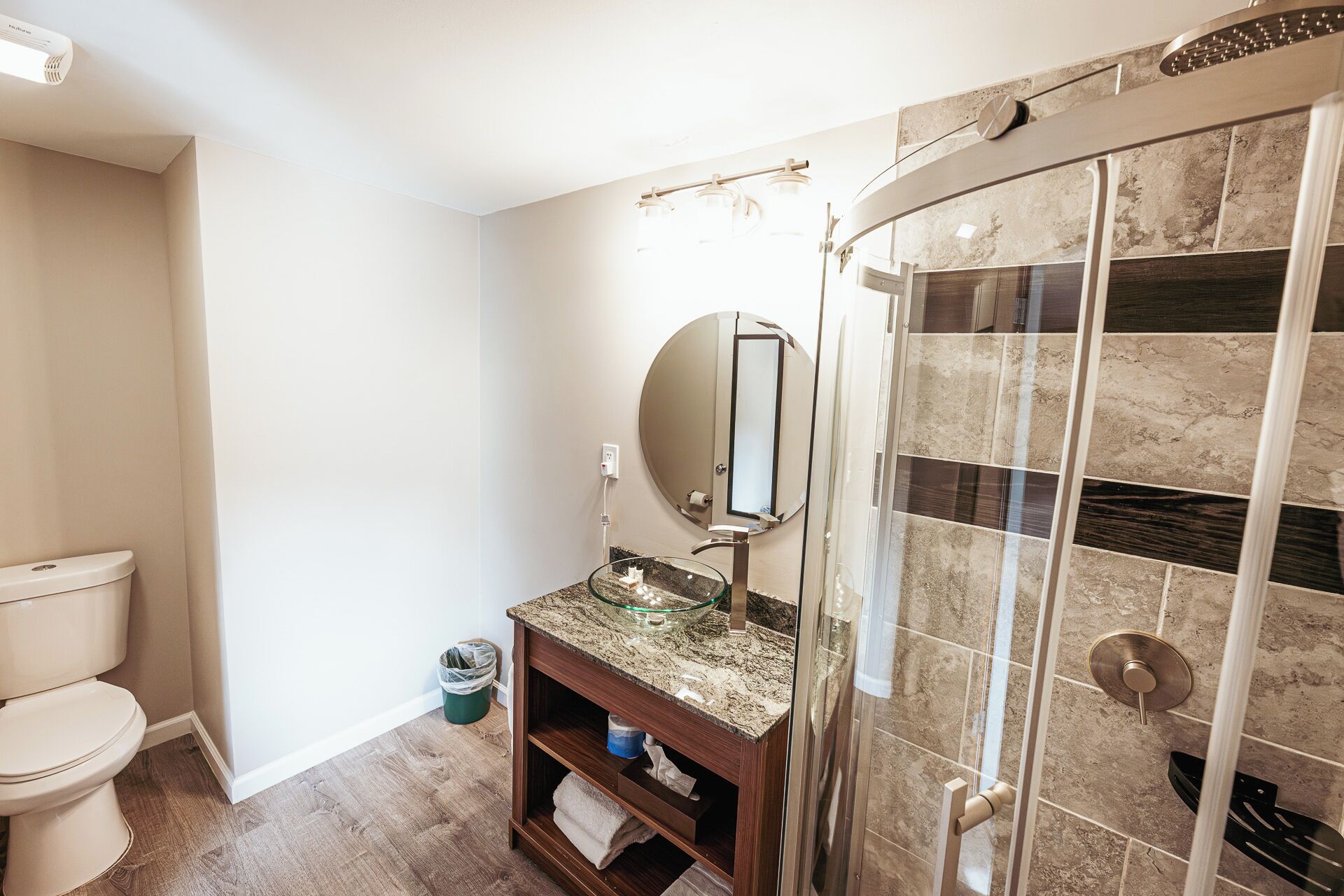 A bathroom with a toilet, a glass sink with a marble countertop, a round mirror, and a shower with a glass door.