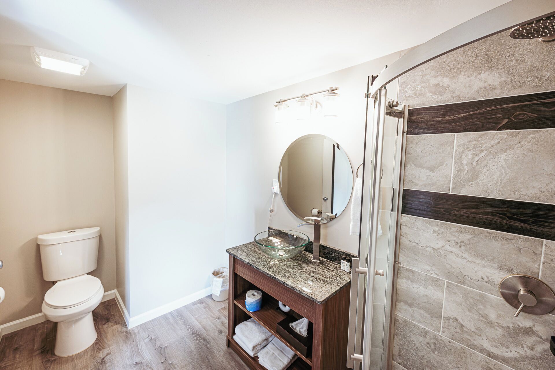 A modern bathroom with a toilet, a glass sink with a marble countertop, a large mirror, and a shower area with a tiled wall.