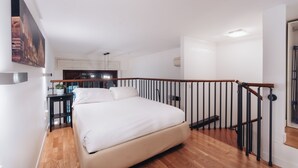Ein Schlafzimmer mit Bett, Holzboden, schwarzem Geländer und Blick zur Küche.