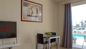 A living room with a flat-screen TV displaying 'NETFLIX', a framed abstract painting, a dining table with chairs, and a view of a swimming pool and cars outside. - Kato Paphos Suites - GF Studio Apartment. Pool View, Wi-Fi, a/c. Great Location (Pafos)