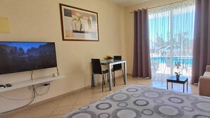 A hotel room with a bed, a desk with two chairs, a TV, a painting, and a view of a pool. - Kato Paphos Suites - GF Studio Apartment. Pool View, Wi-Fi, a/c. Great Location (Pafos)
