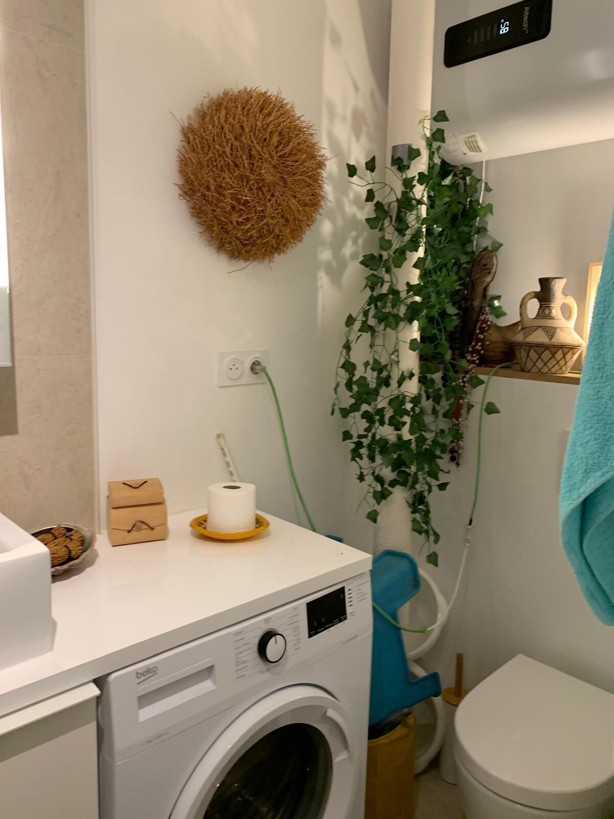 Une petite salle de bain équipée d’un lave-linge, de toilettes et d’une étagère où sont disposés des objets décoratifs.
