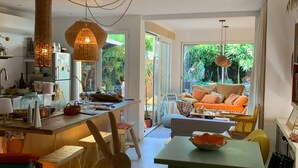 A modern kitchen with a dining area, featuring a large window, wicker pendant lights, and a cozy seating area with cushions. - Large studio with garden: a haven of freshness and greenery right in the center of town (Cavalaire-sur-Mer)
