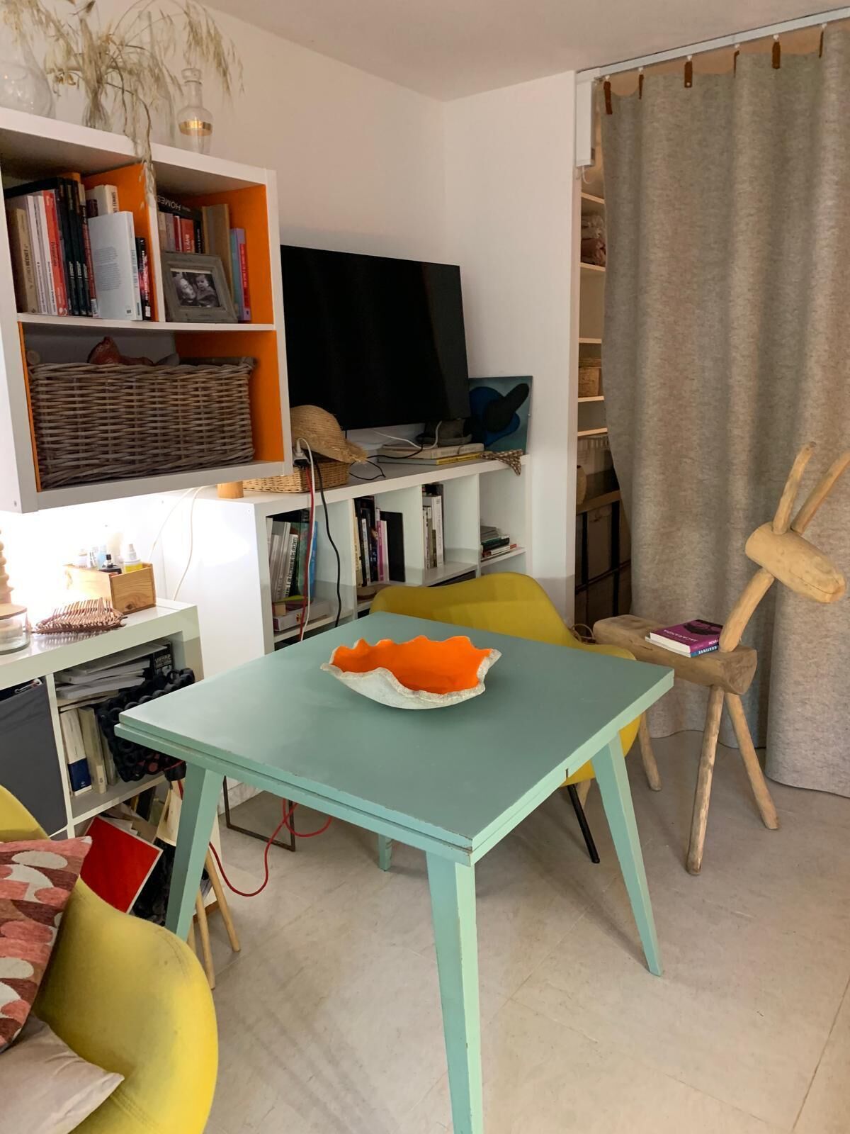 Ein Zimmer mit einem Türkis-farbenen Tisch, gelben Stühlen, einem Bücherregal und einem Bild an der Wand.