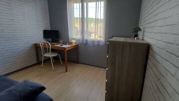 Una habitación con escritorio, silla y televisión. Hay una ventana con cortinas, una planta en un estante y un sofá.