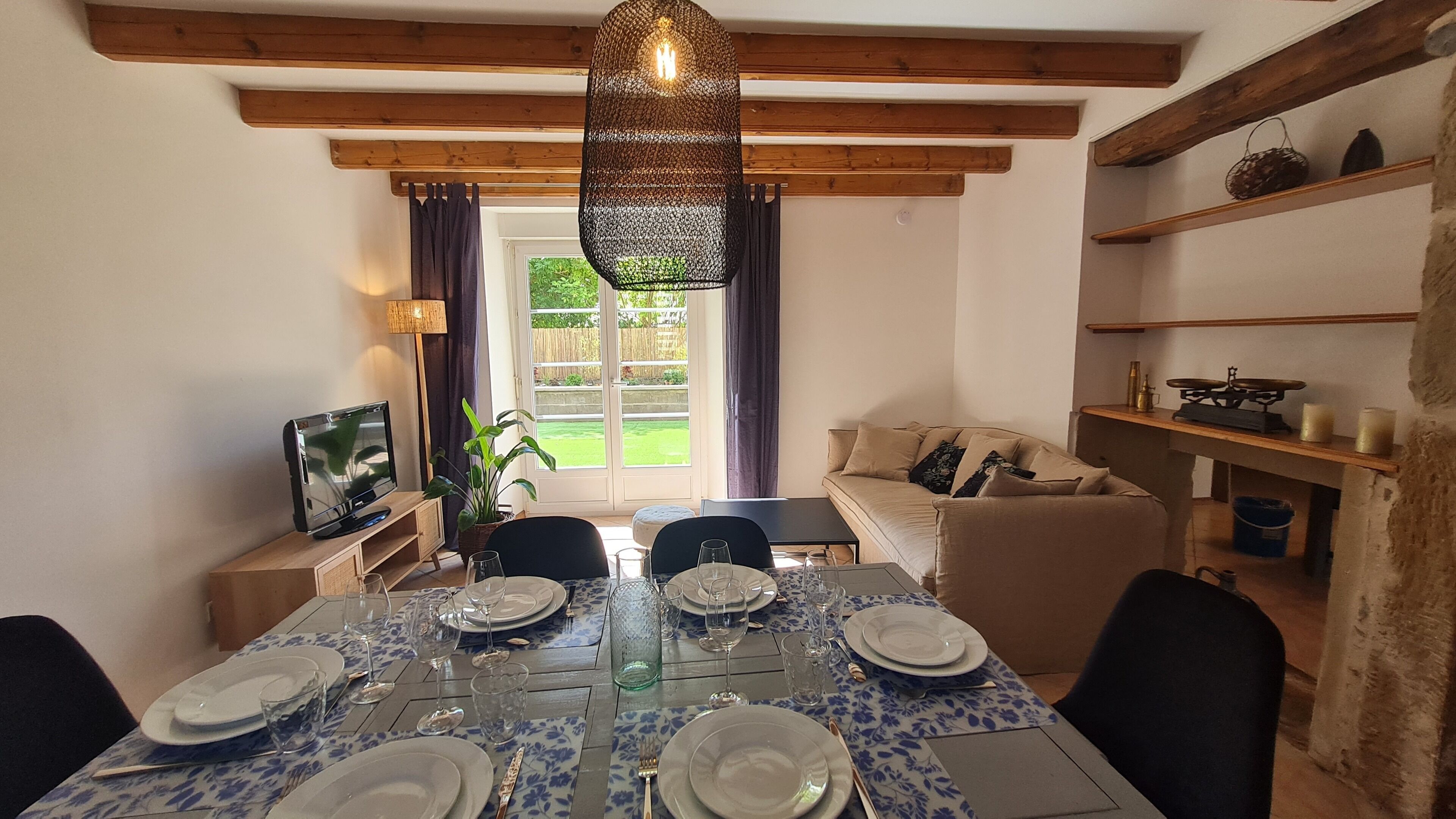 Un comedor con una mesa dispuesta para seis personas, un televisor sobre un atril de madera, y vistas al jardín a través de la ventana.