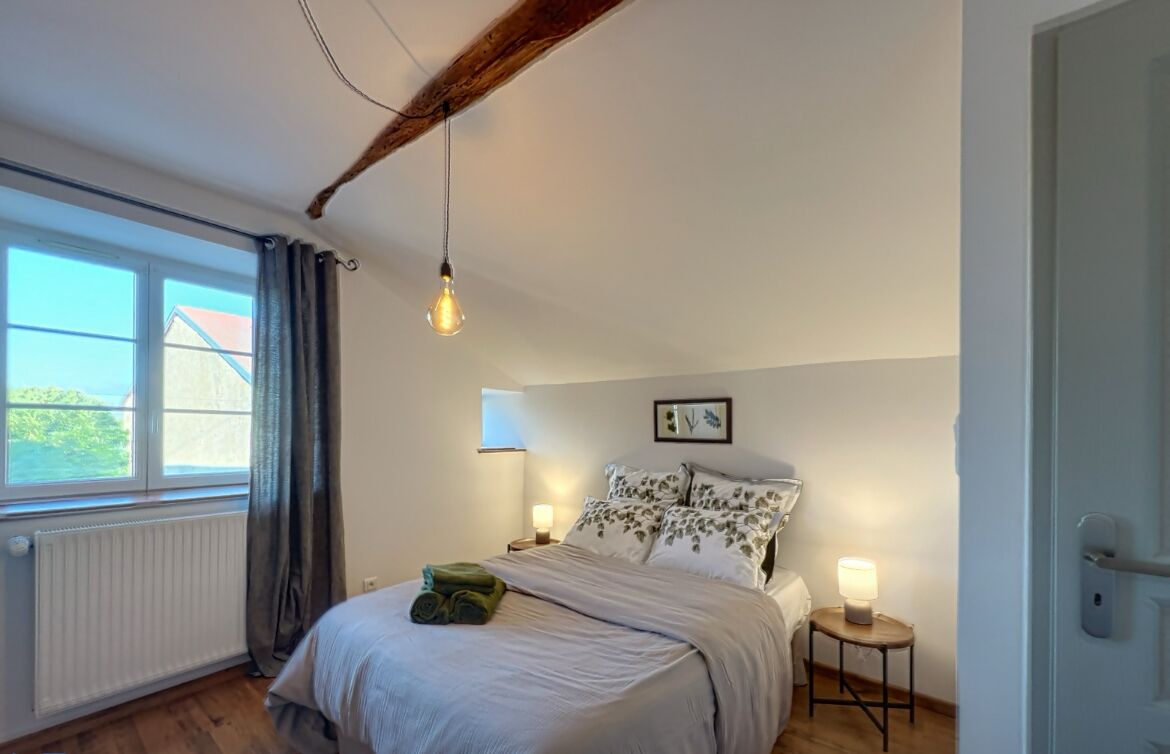 Een slaapkamer met een bed, nachtlampjes, een raam met gordijnen, een radiator en een houten balk in het plafond.