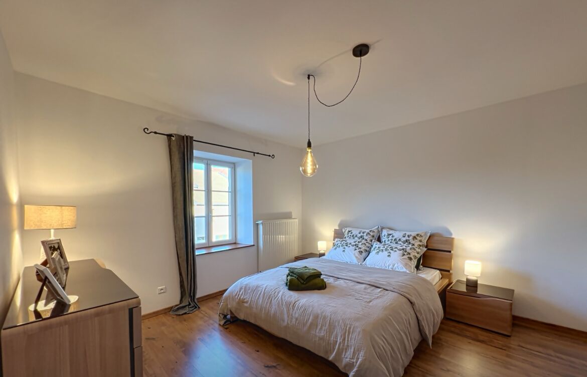 Quarto com cama, mesas de cabeceira, luminária, radiador, janela com cortinas e luminária pendente.