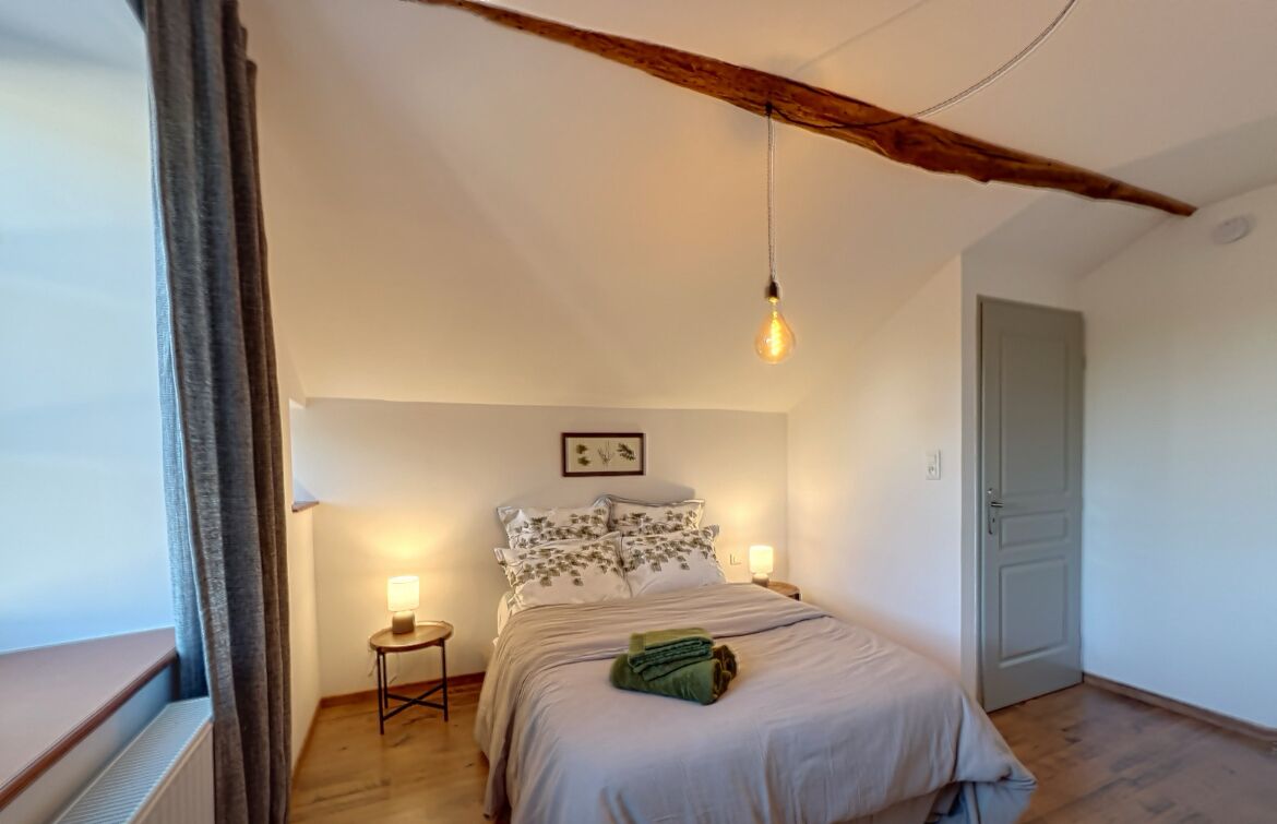A bedroom with a bed, bedside lamps, a window with curtains, and a wooden ceiling beam.