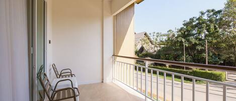 A balcony with a table and chairs, a view of a road and greenery, and a building in the background.