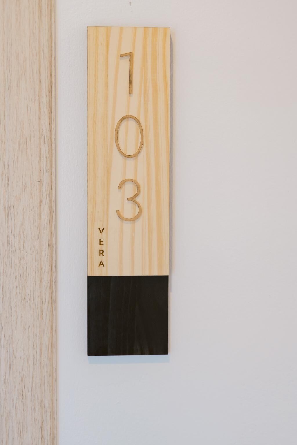 Une plaque en bois portant les chiffres « 703 » et le mot « VERA », fixée à un mur blanc.