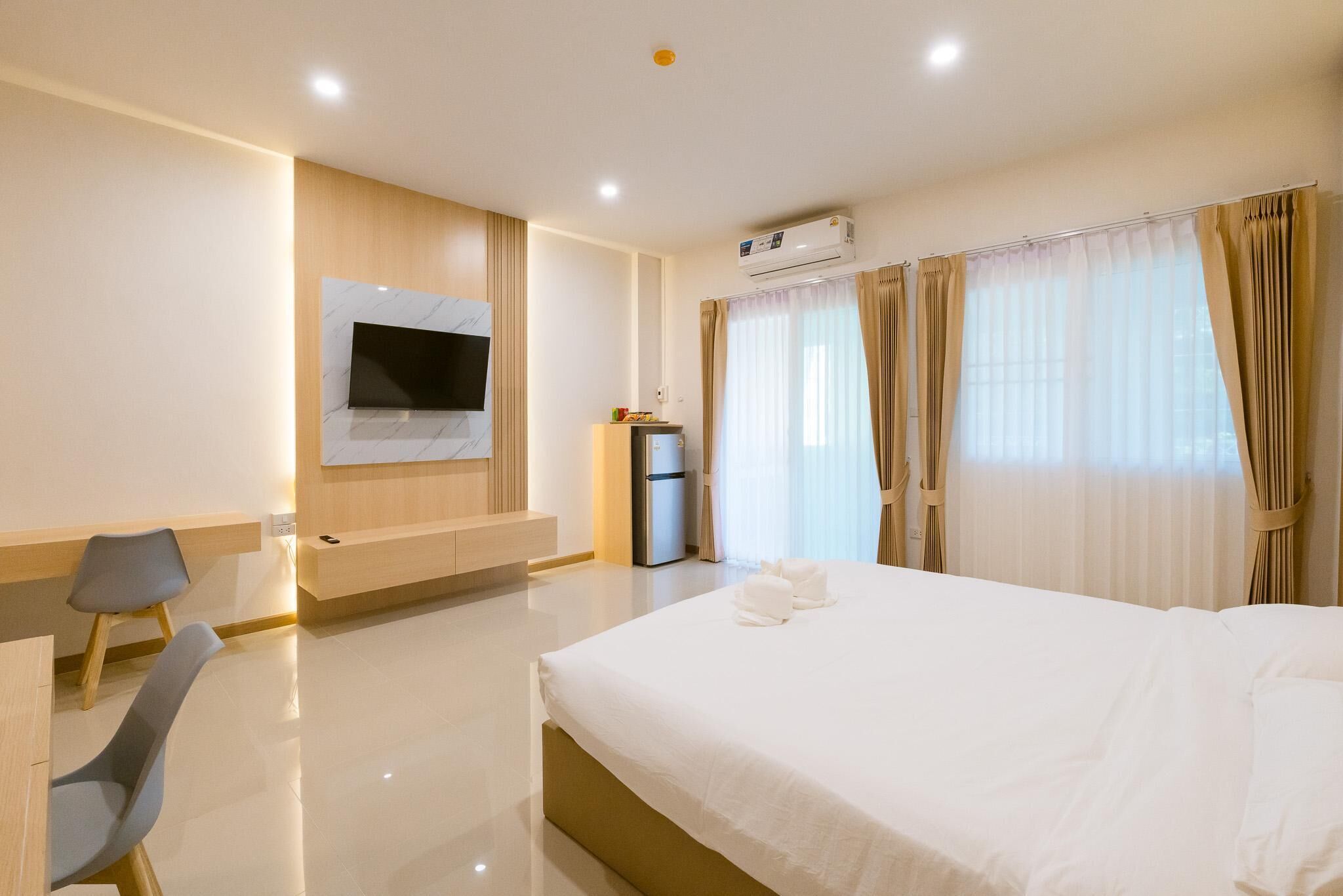 A modern hotel room with a large bed, a flat-screen TV mounted on the wall, a desk with a chair, and a minibar.