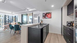 A modern kitchen with a central island, a dining area with chairs, and a large window offering a city view. - South Bank Sensational! ~ 2Bed/2Bath/1Car/Pool! (South Brisbane)