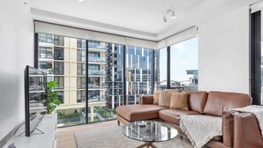 A modern living room with a large window, a leather sofa, a glass coffee table, and a flat-screen TV. - South Bank Sensational! ~ 2Bed/2Bath/1Car/Pool! (South Brisbane)