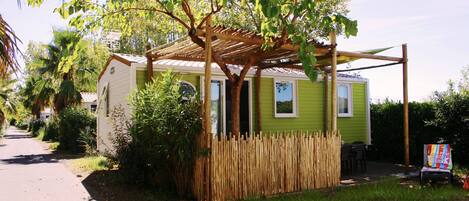 Une petite maison de plain-pied, à l’extérieur vert, avec une clôture en bois et une structure en forme de pergola.