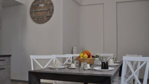 A dining area with a table set for four, white chairs, a fruit bowl, and a wooden clock on the wall.