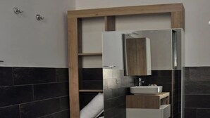 A modern bathroom with dark tiles, a wooden-framed mirror, a white sink, and a built-in shelf with towels.