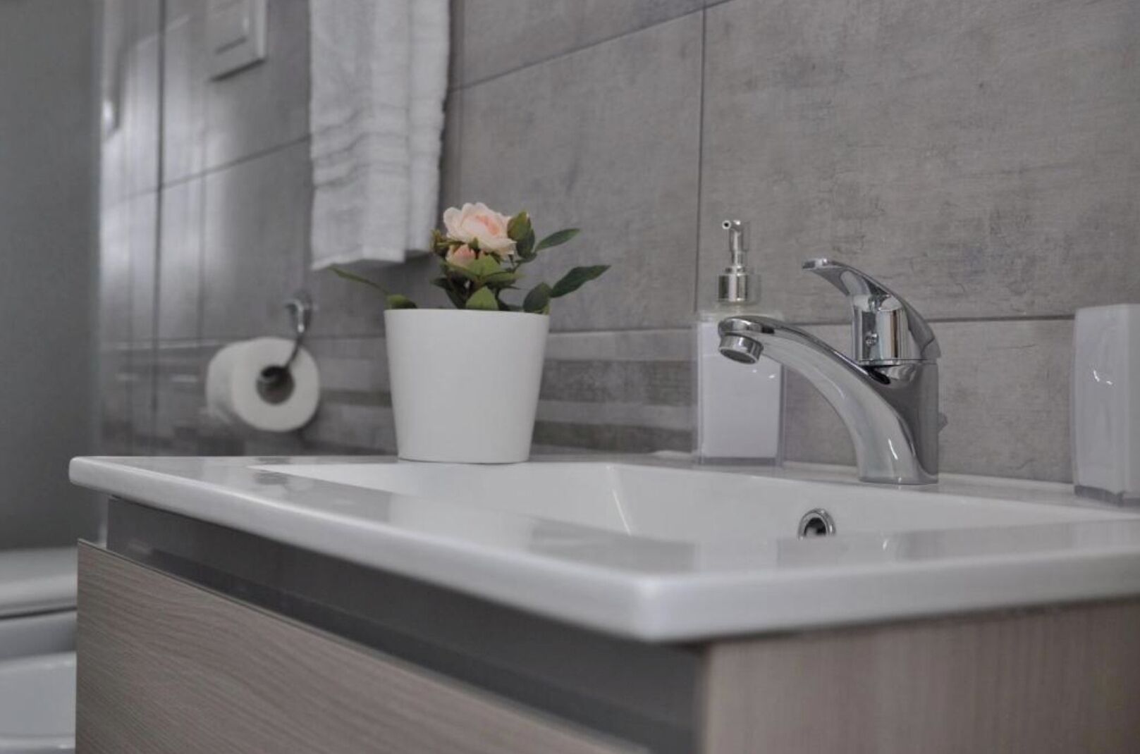 Et badeværelse med en hvid vask, en moderne vandhane, en potteplante og en toiletpapirholder.
