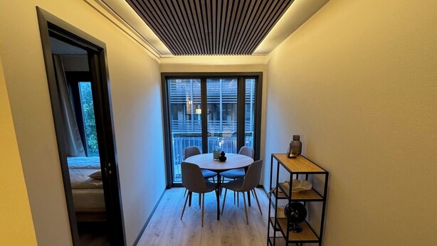 A narrow hallway with a wooden floor, a small table with chairs, and a window with a view of buildings. - 2-bedroom apartment in Fredrikstad (Østfold)