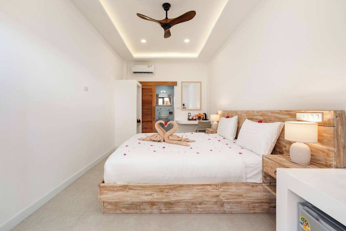 Camera d'albergo moderna con una struttura letto in legno, una ventola a soffitto, un comodino con una lampada e vista sul bagno.
