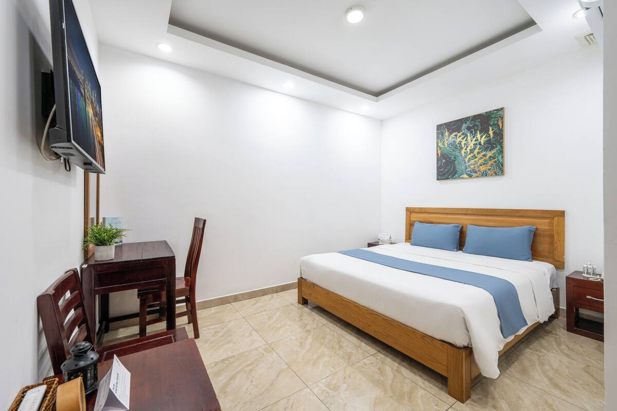 A modern hotel room with a bed, bedside table, and a desk with a chair.
