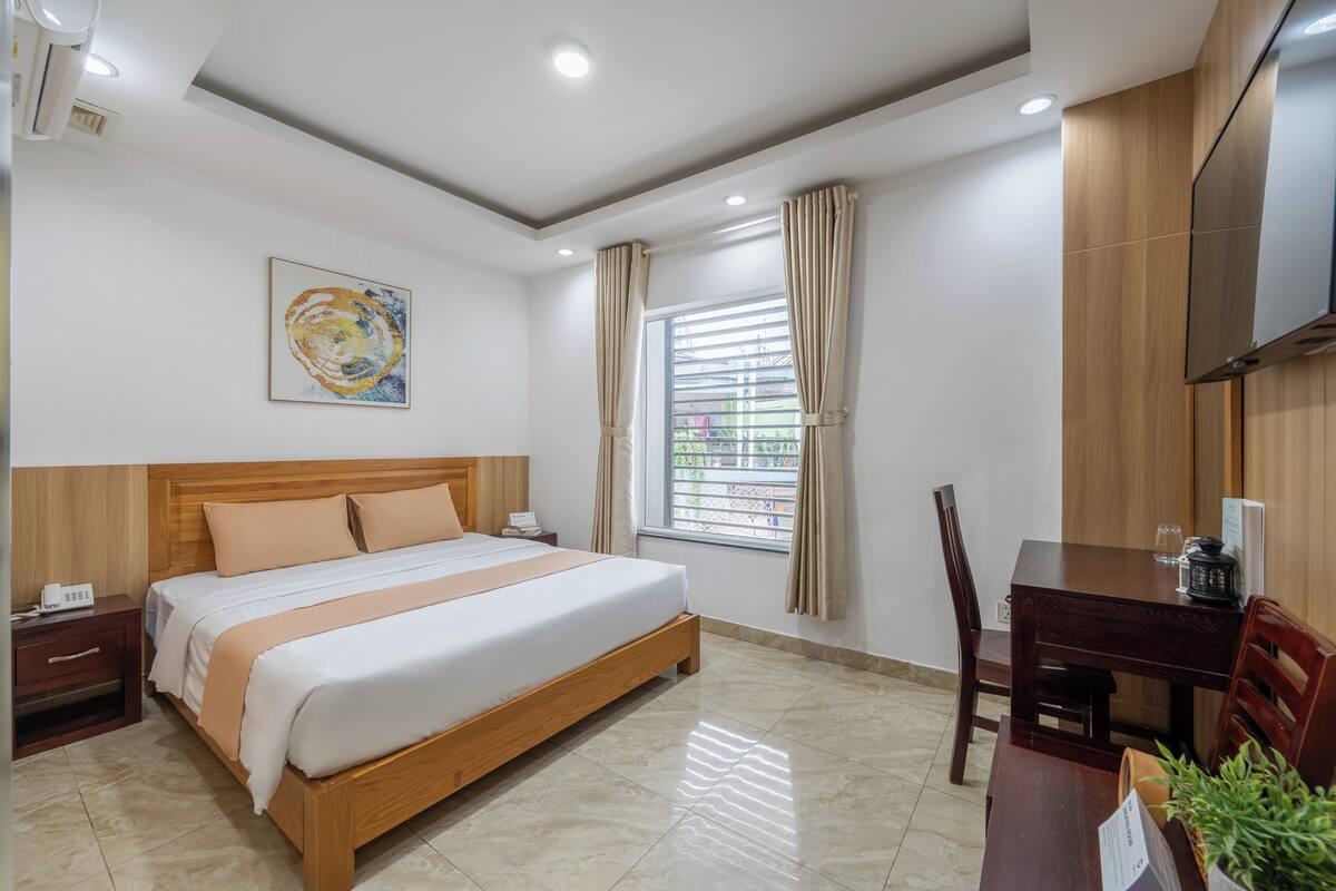 A hotel room with a wooden bed, a desk with a chair, a TV mounted on the wall, and a window with curtains.