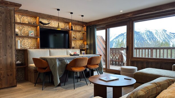 Un comedor acogedor con una mesa y sillas de madera, un televisor sobre la encimera, y vistas a montañas a través de amplios ventanales.