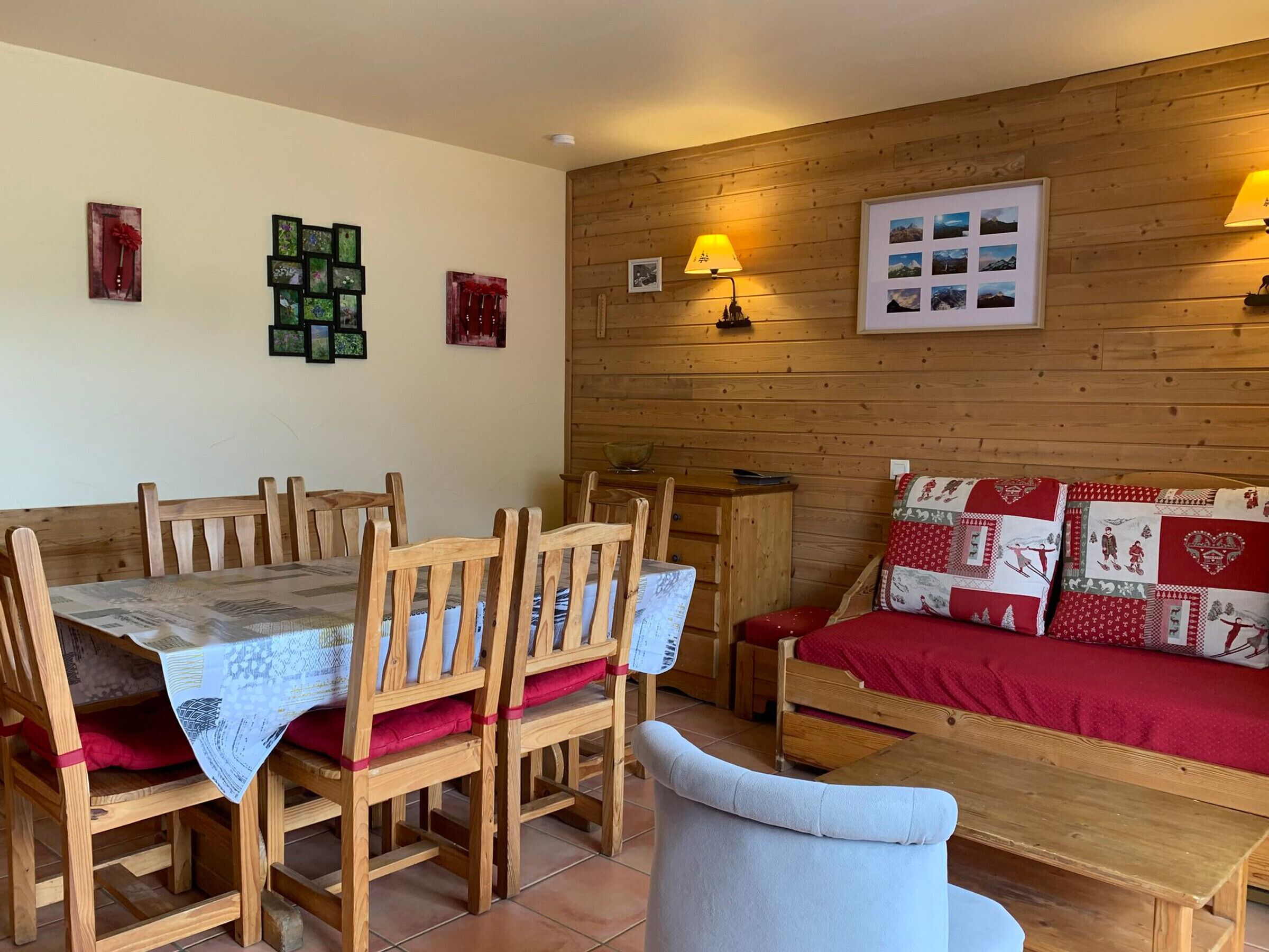 Ein gemütliches Zimmer mit einem Holzesstisch und -stühlen, einem roten Sofa mit gemusterten Kissen und einer Wand aus Holz mit gerahmten Bildern.